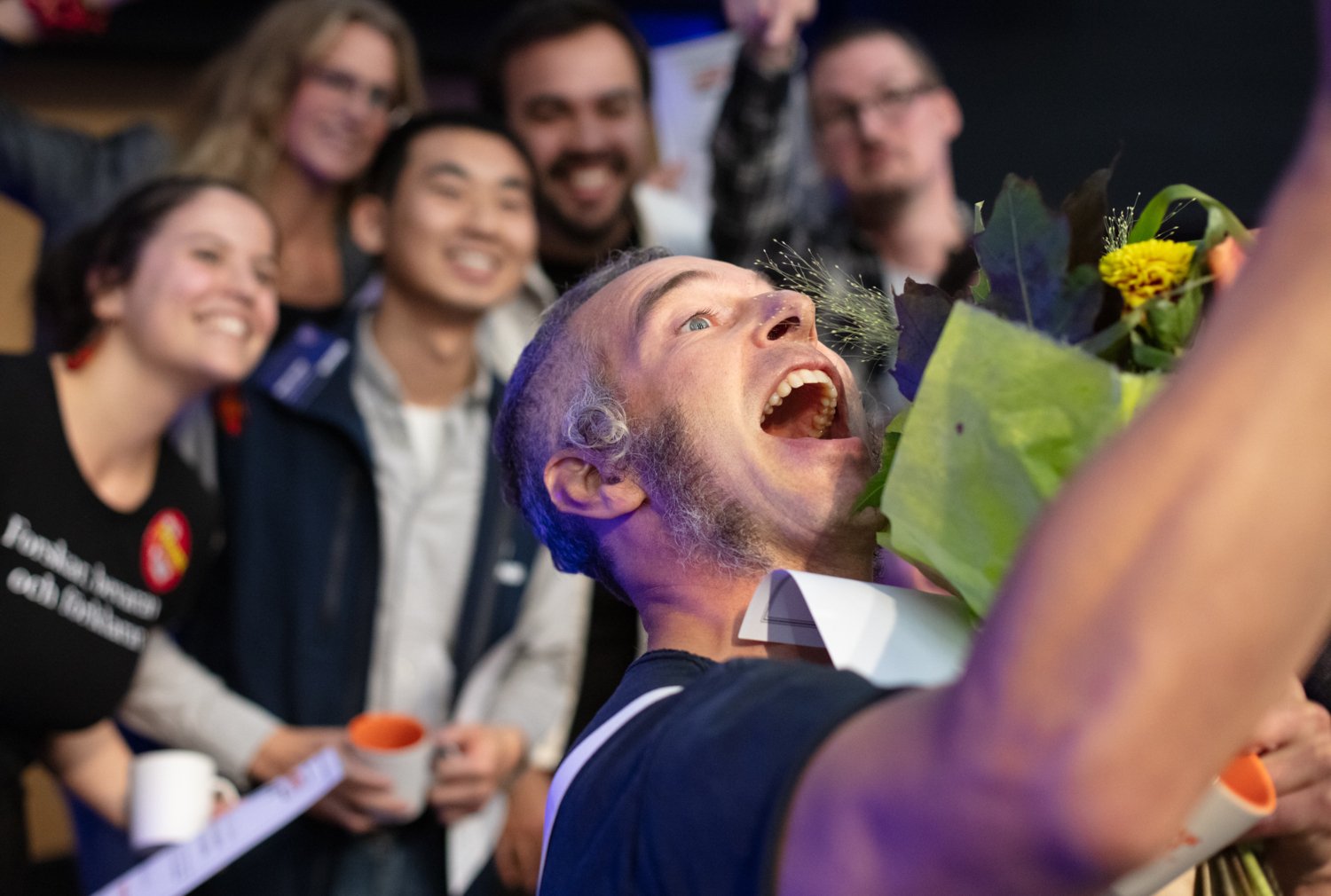 Glad man tar en selfie tillsammans med fem personer i bakgrunden vars ansikten är lite suddiga.