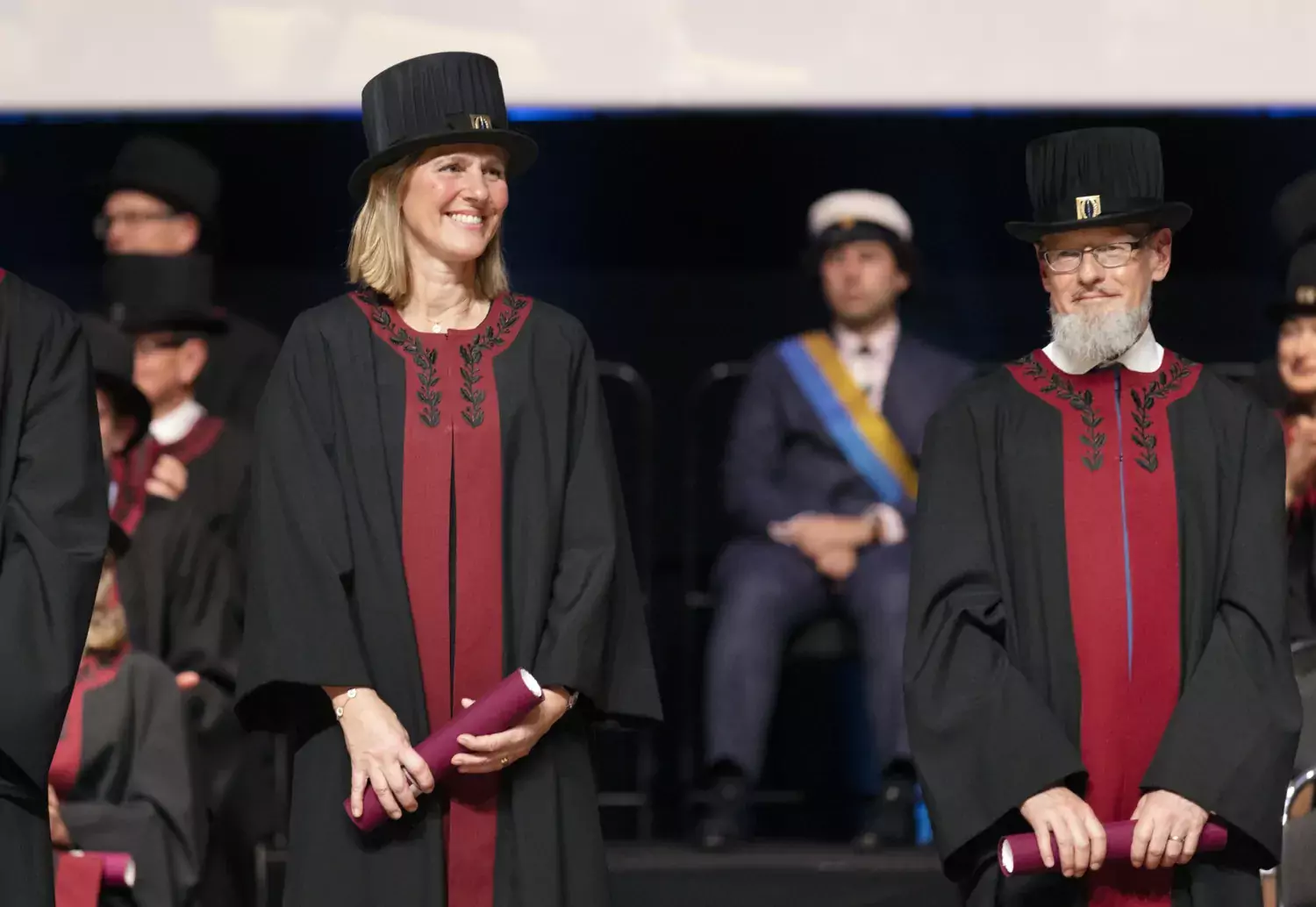 Kristina Haugaa i förgrunden, håller i sitt diplom och ler. Lars E Eriksson står i bakgrunden.