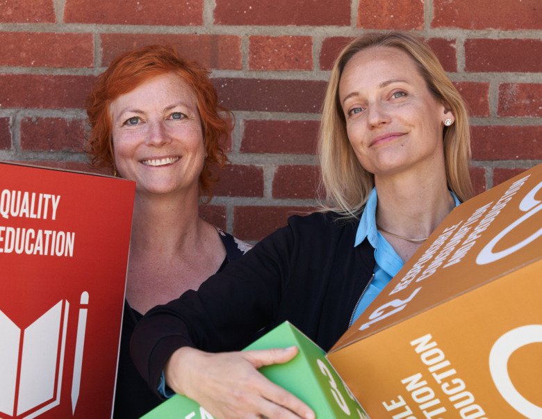 Sara Widén och Hanna Karlsson står med kuber som föreställer tre av de globala hållbarhetsmålen framför en tegelvägg.