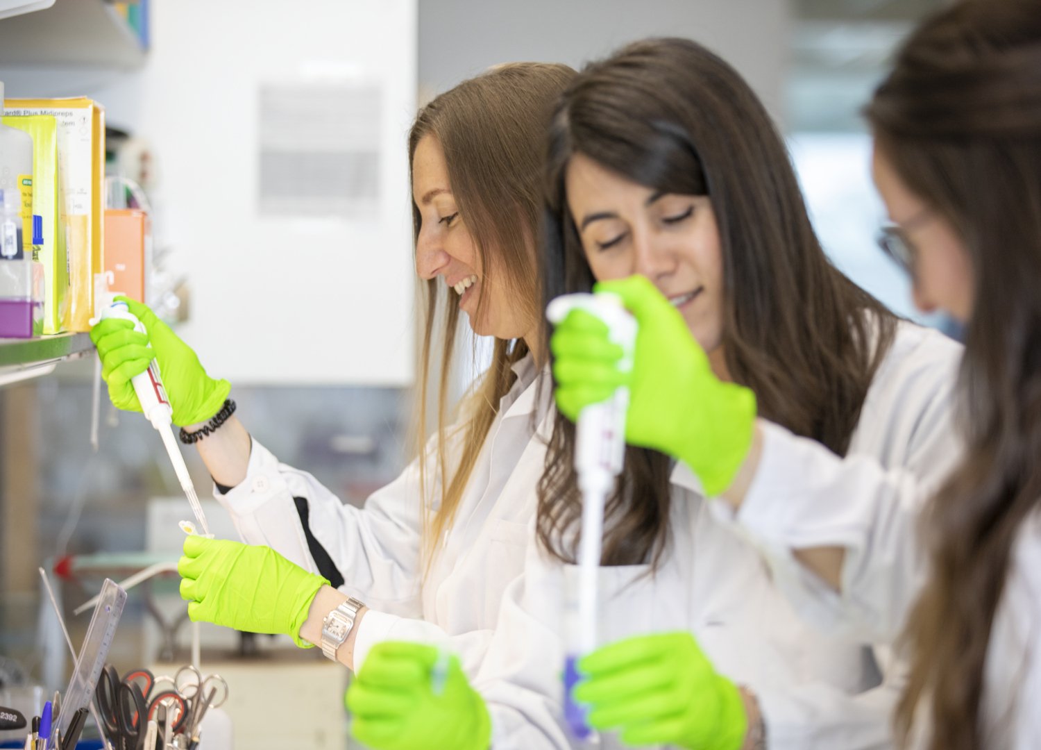 Tre kvinnliga forskare står med provrör och pipetter i labbmiljö.