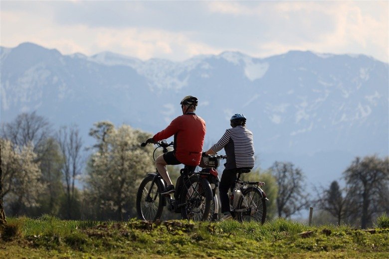 Bild av två äldre personer som cyklar i naturen, troligen tidig vår, båda har hjälm på sig.