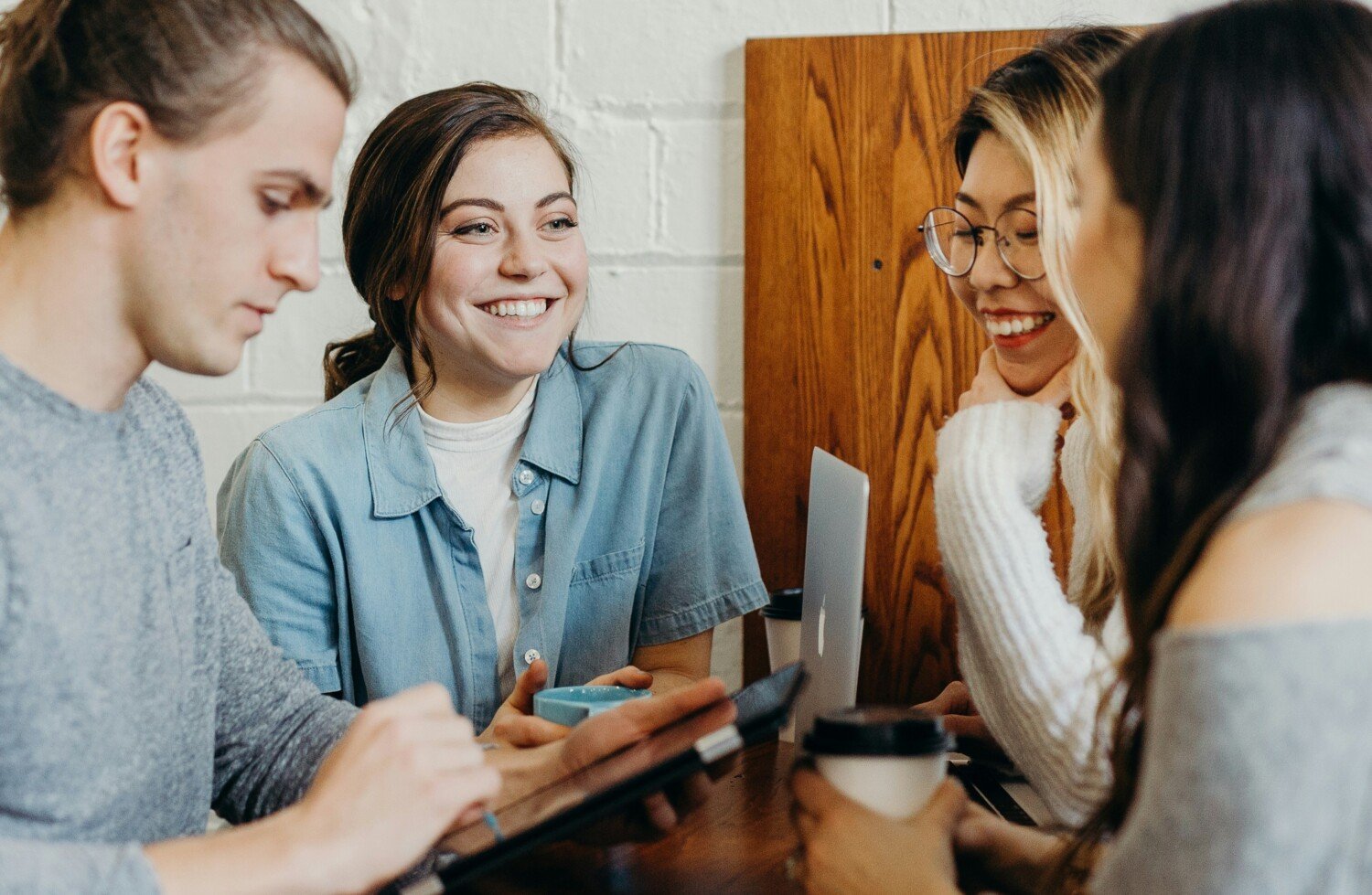 En grupp med fyra yngre personer sitter runt ett bord och ler mot varandra, kaffemugg i handen på en av dem