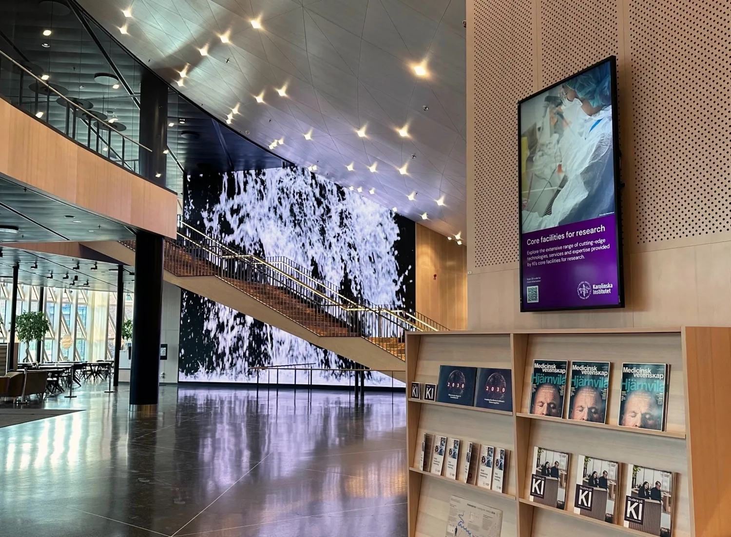 Digital skärm intill reception ovanför hylla med tidskrifter och annat tryckmaterial. I bakgrunden skymtar bildspel på väggen och trappan upp till plan 3.