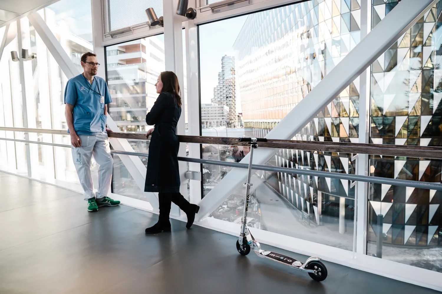 Man i sjukhuspersonalkläder som jobbar på Karolinska universitetssjukhuset samtalar med kvinna i svarta kläder. De ståri gången som leder mellan KI och sjukhuset.