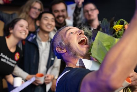 Glad man tar en selfie tillsammans med fem personer i bakgrunden vars ansikten är lite suddiga.