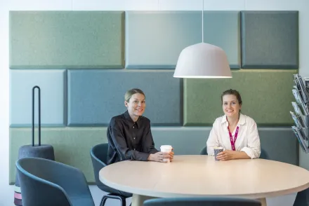 Två kvinnor sitter vid ett bord med varsin kaffe i handen och tittar in i kameran.