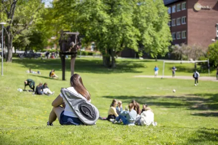 Kvinna med badmintonrackväska på ryggen sitter på gräsmatta på campus Solna, med personer i bakgrunden.